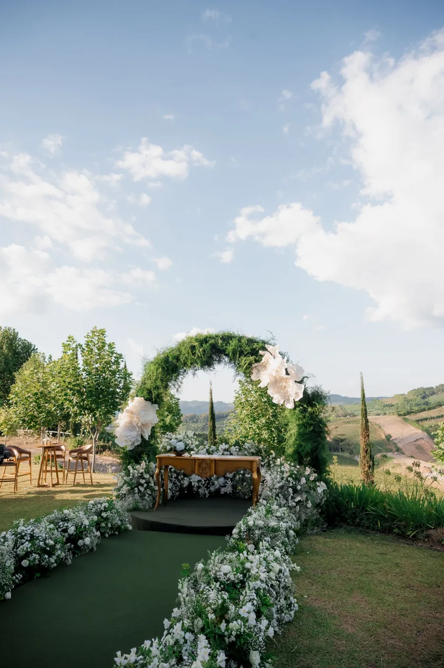A round floral arch with white blooms and greenery on a hillside, with aisle florals leading to it