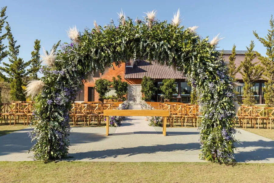 A wide greenery arch on a lawn with chairs arranged for a ceremony
