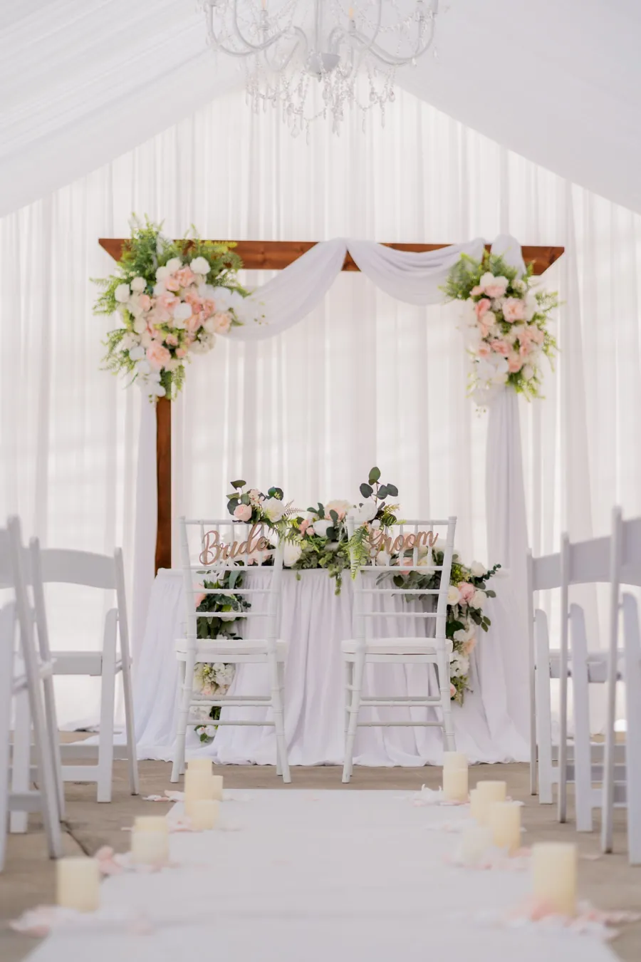 A wooden ceremony arch with blush and ivory florals at the corners, draped in white, inside a tented ceremony