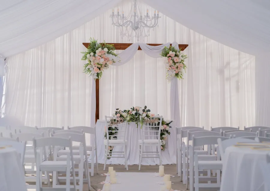 A tented ceremony with a wooden arch, blush florals and white chairs
